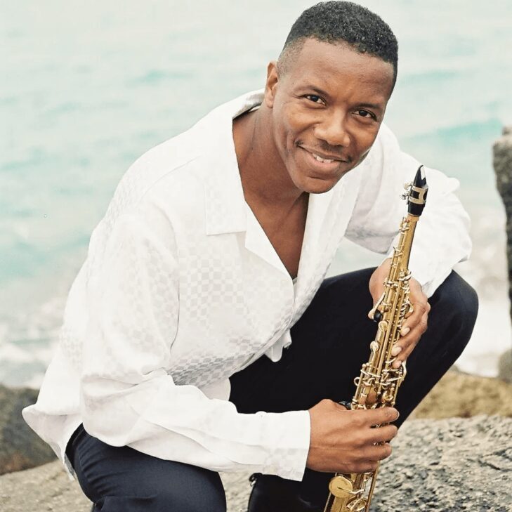 A man is posing with a saxophone in front of the ocean.